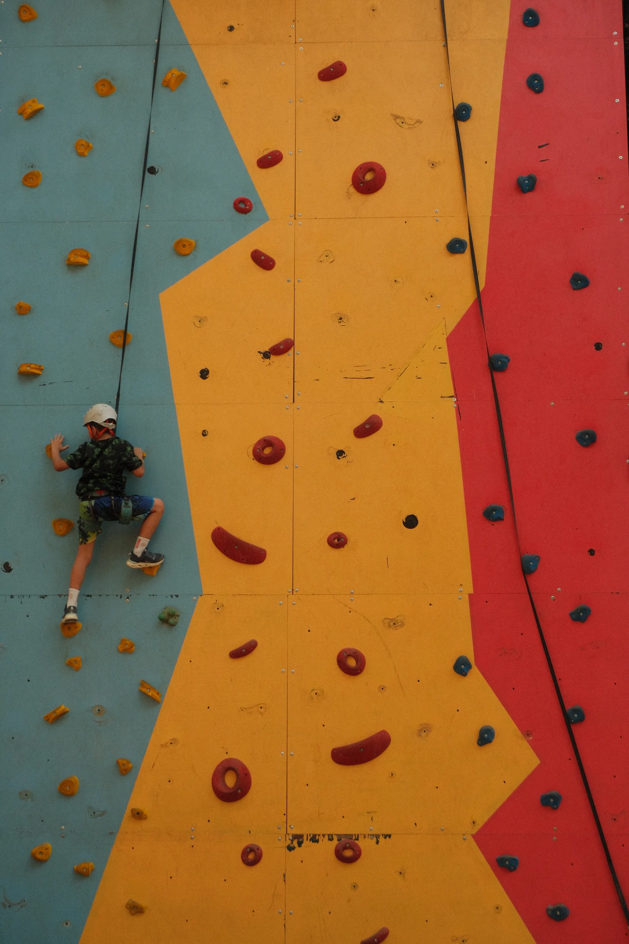 A child in a helmet climbing a color-blocked indoor climbing wall.